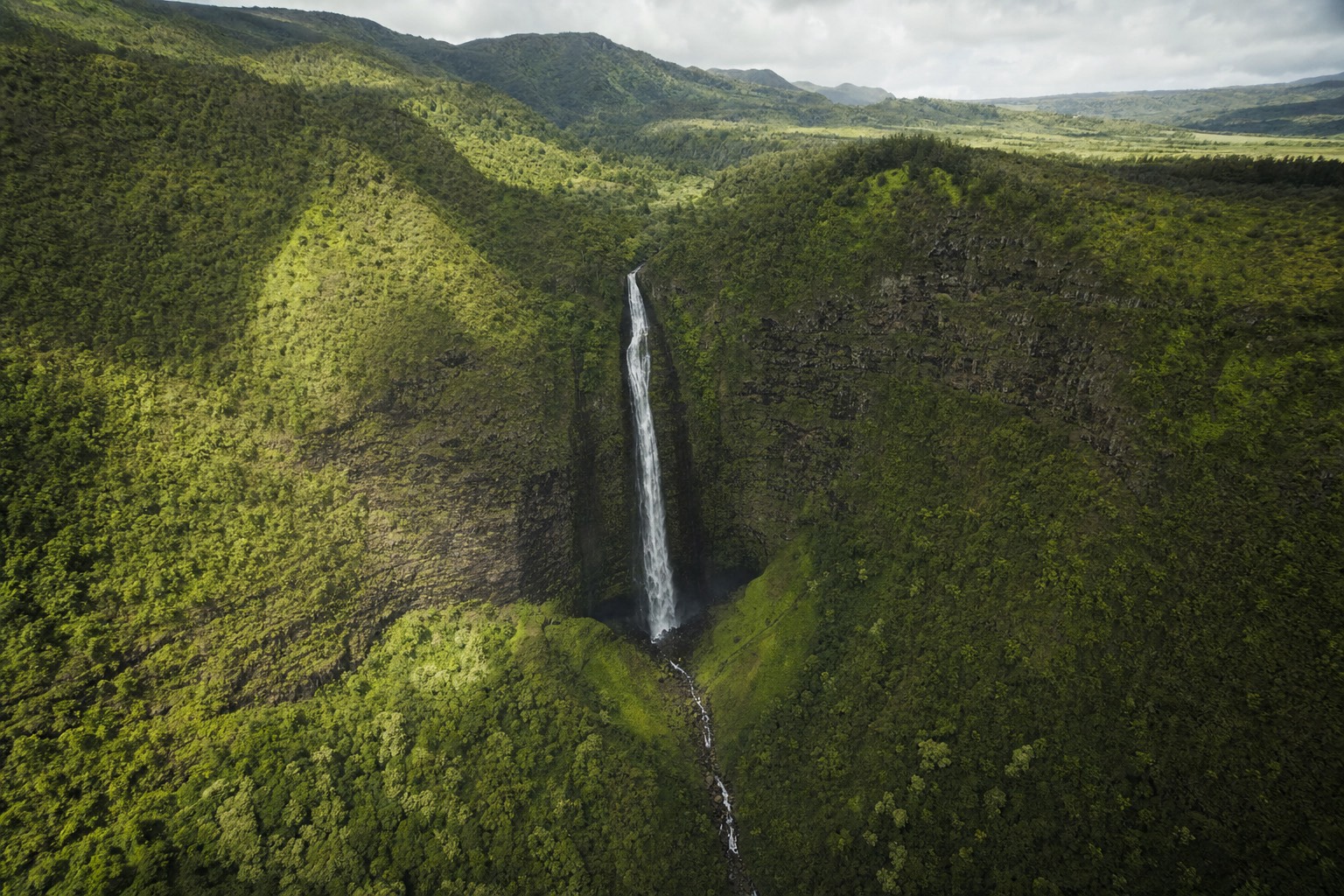 Helicopter view of a tall waterfall dropping through green jungle cliffs in a Kauai valley