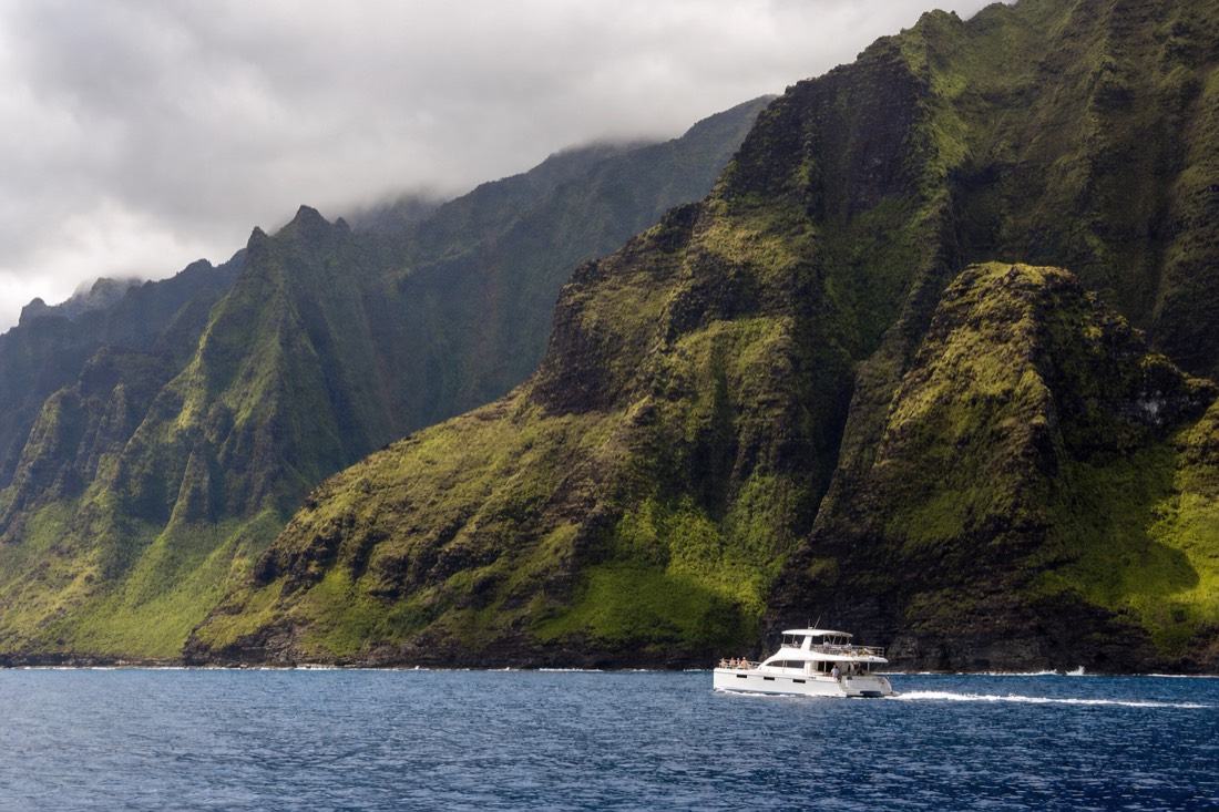 A catamaran cruising beneath the Na Pali Coast cliffs on Kauai, the flagship boat tour in these Kauai tour packages